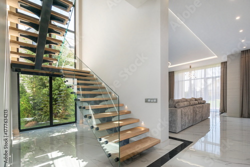 An elegant wooden staircase with glass railings leads to a spacious living room filled with natural light.