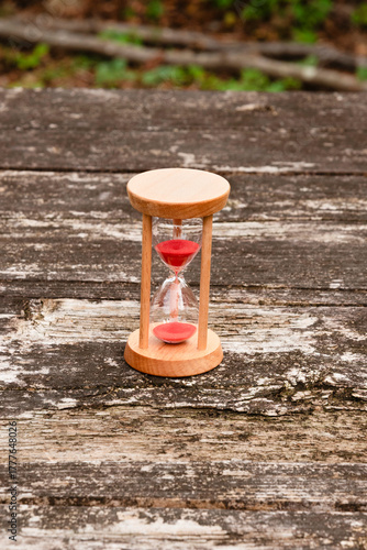 Wooden hourglass with red sand running through, standing on a textured, weathered rustic wooden table. Outdoor setting. Symbolizes time, passage, countdown, and deadline.