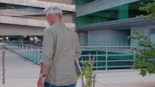 Senior businessman with gray hair walking with laptop in hand outdoors in business district. Wearing smart casual outfit, he appears focused and confident in daylight setting
