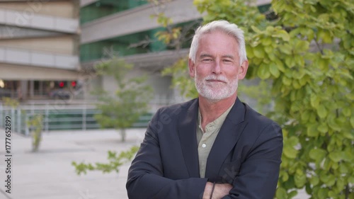 Zoom in shot of confident senior businessman with white hair and beard standing outdoors near modern office with arms crossed, showing leadership and success in corporate world