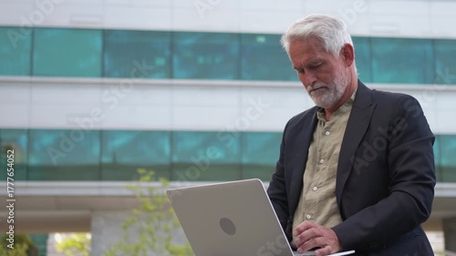 Confident senior businessman using laptop outdoors near office building, working remotely in modern urban environment during daytime