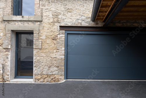 Foto Porte de garage grise anthracite installée par un professionnel, encadrée par un mur en pierre beige et un sol en macadam, entre modernité et authenticité
