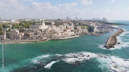 Aerial view of Old Jaffa's sandstone buildings meeting the turquoise Mediterranean Sea, a vibrant contrast against the distant Tel Aviv skyline, Old Jaffa, Tel Aviv-Yafo, Israel.