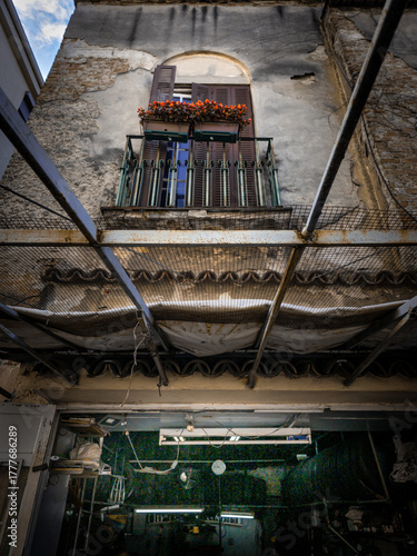 Wallpaper Mural A glimpse up a weathered Belgrade building, where faded walls and rusted beams frame a small balcony adorned with red flowers and closed shutters, . Torontodigital.ca
