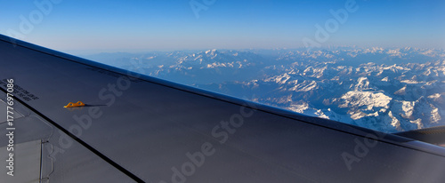 View from an airplane window, with a wing in the foreground and a snow-capped mountain range on the horizon. The mountain range depicted is the Alps, seen from cruising altitude.