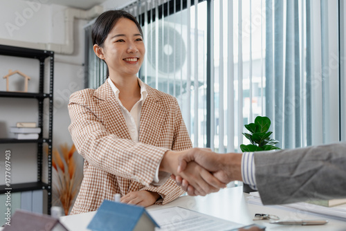 Two people in business attire finalize a real estate deal, shaking hands across a desk with house models, keys, and a contract, symbolizing agreement, trust, and successful negotiation.