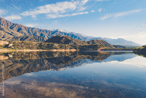 Natural landscape of mountains perfectly reflected in a crystal-clear lake under a clear sky. The scene conveys calm and purity, with the golden tones of the sun highlighting the textures of the moun