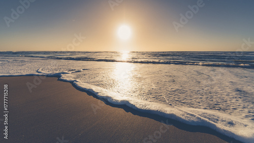 The image shows a beach at sunrise. In the foreground, you can see the wet sand and the foam from a wave that has just broken gently on the shore. The sea occupies much of the scene, with small waves