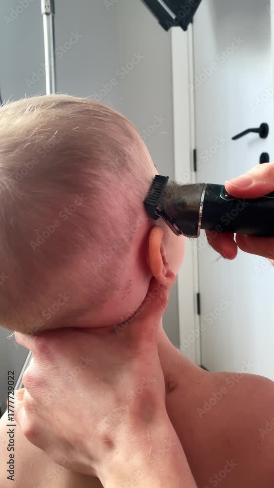 A a toddler boy getting his hair trimmed skillfully mother cuts her little boy's hair at home, using a clipper for a neat and trendy look. Mother hold with hand the neck while she works.