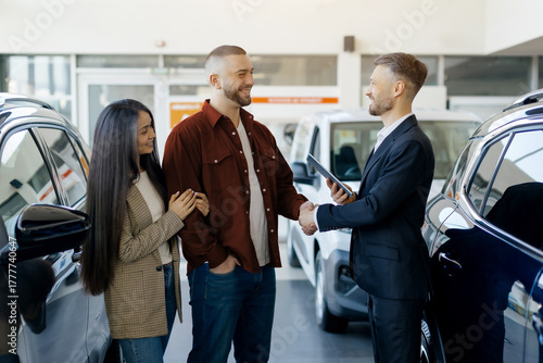 Friendly salesman shaking hands with a smiling couple inside a bright, modern car dealership center, cars visible around them. Happy young family buying new vehicle in auto salon