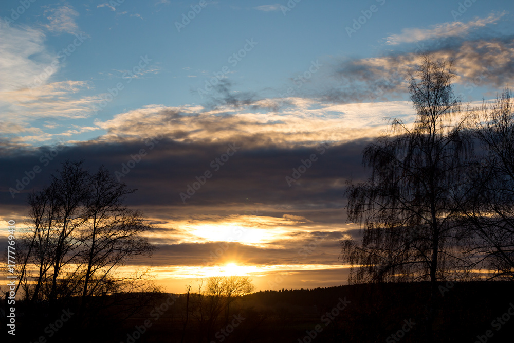 Fototapeta premium Radiant sun peeks through dramatic clouds, painting the autumn sky with fiery orange and gold. Leafless trees create striking silhouettes against the vivid sunset vista