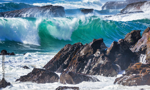 Powerful turquoise ocean waves crashing against rugged coastal rocks with white sea foam and splashing spray under clear blue sky showing the raw beauty and unstoppable energy of the ocean nature