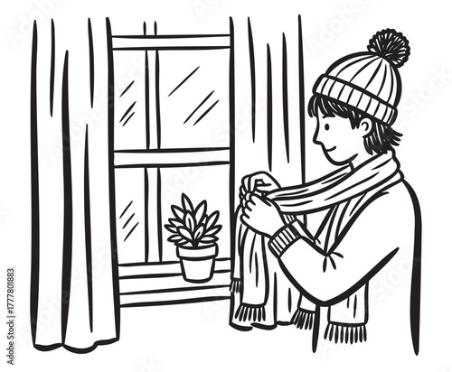 Young caucasian male wearing a winter hat and scarf stands near a window with curtains, adjusting his scarf, next to a small potted plant.