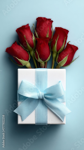 Elegant bouquet of fresh red roses with lush green leaves arranged behind a beautifully wrapped gift box adorned with a pastel blue satin ribbon on a soft blue background
