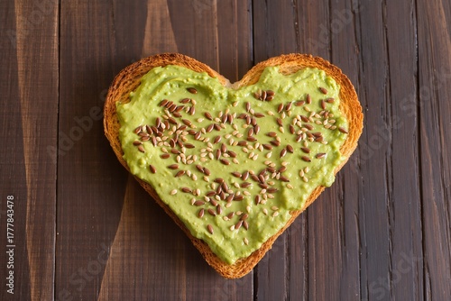 Heart-shaped toasted bread spread with avocado paste and sprinkled with flax and sesame seeds on a dark wooden background, top view