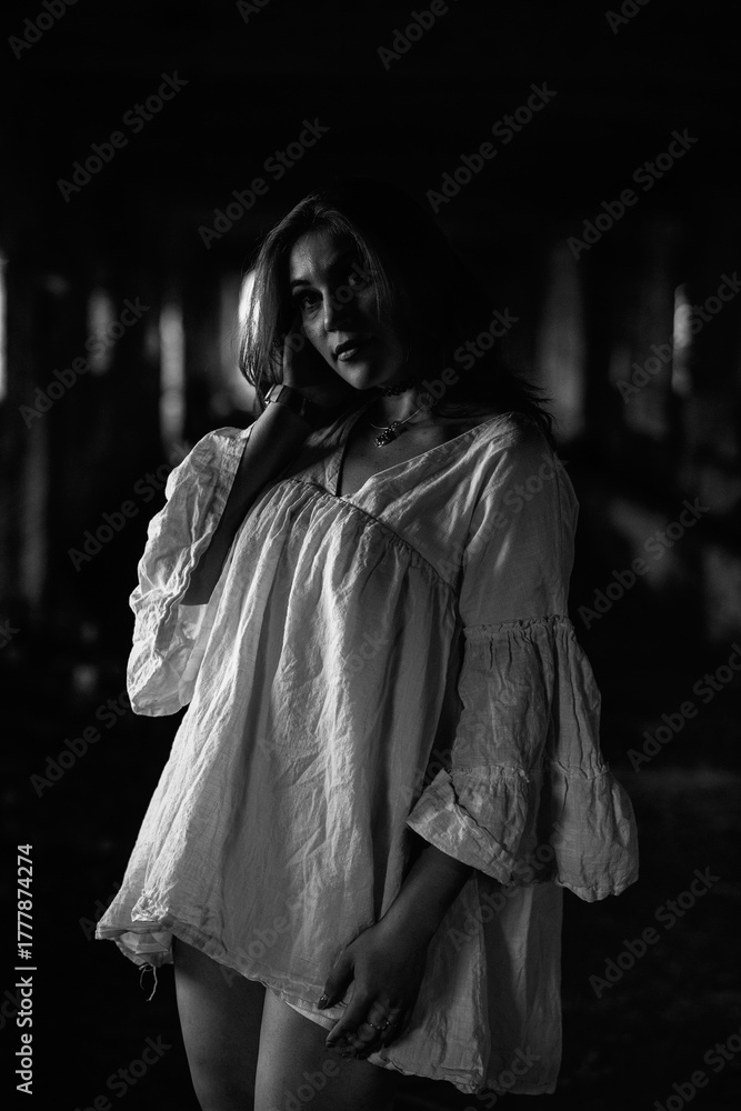 Naklejka premium Young woman with long hair wearing a white oversized shirt poses confidently in an abandoned building, showcasing a blend of elegance and urban decay in monochrome