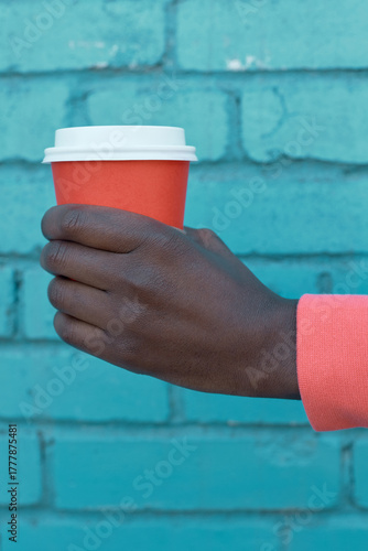 Close-up of Black person's hand in coral sleeve holding orange takeaway cup with white lid against vibrant blue brick wall creating bold urban contrast