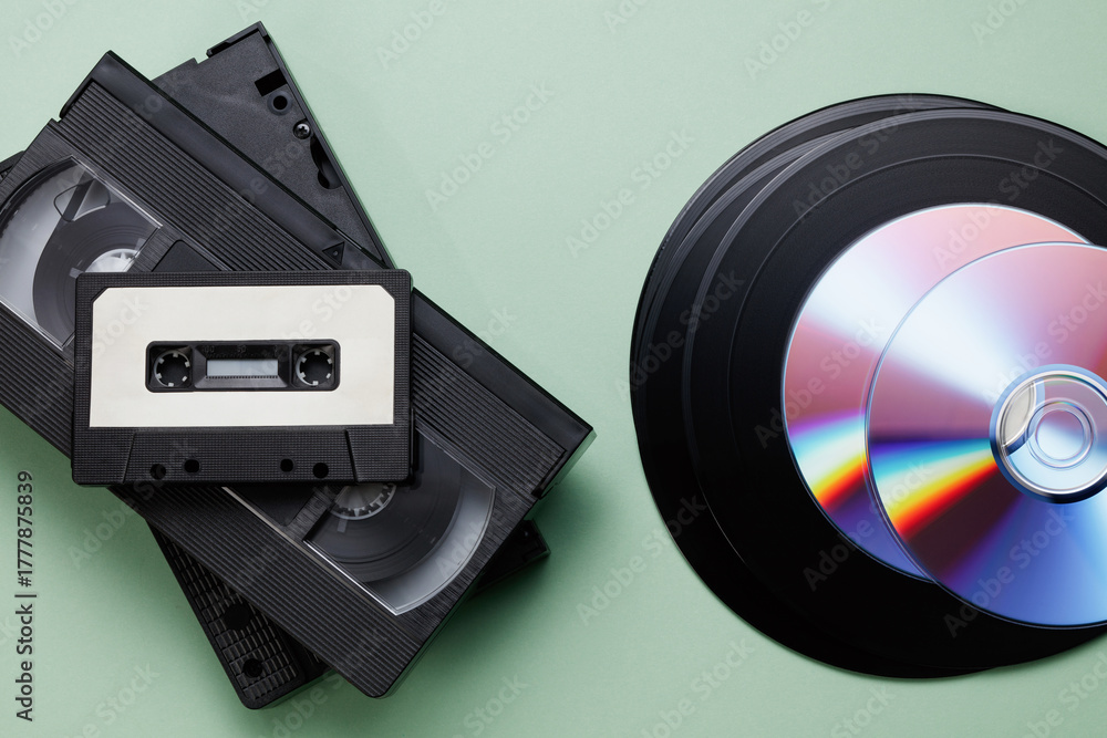 Obraz premium Texture of vintage retro vinyl records, retro acoustic, audio cassette tape, black video tape cassette, CD disc on a green background. Saving data, music, movies, videos. Top view. 