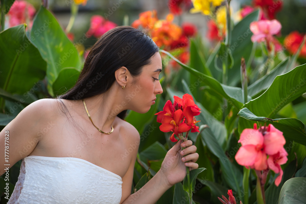 Fototapeta premium Young woman, beautiful, brunette, blue eyes, Hispanic, wearing a white top, surrounded by plants, smelling the scent of beautiful colorful flowers. Concept: model, beauty, fashion, trendy.