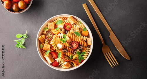 Warm salad with colorful full grain fusilli pasta, olives, fried tofu, cherry tomatoes and aromatic oregano leaves in ceramic bowl on black stone table top view. Healthy vegan food concept.