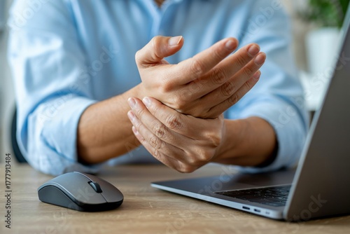 A person is seen holding their wrist, indicating pain or discomfort, while sitting at a desk with a laptop and mouse. 