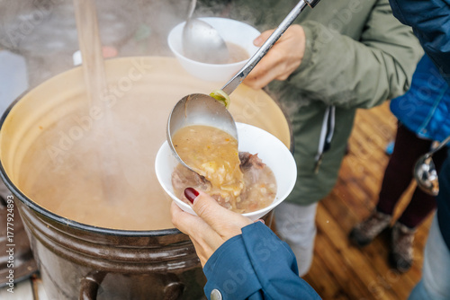 A steaming bowl of hearty soup is served from a large pot outdoors, offering warmth and comfort during a cozy autumn gathering.