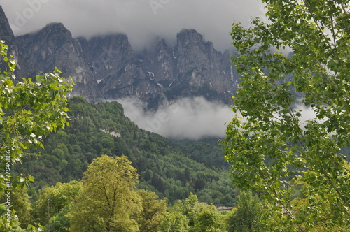 Clouds hang over a serene mountain landscape in the early morning light, revealing lush greenery and rocky peaks
