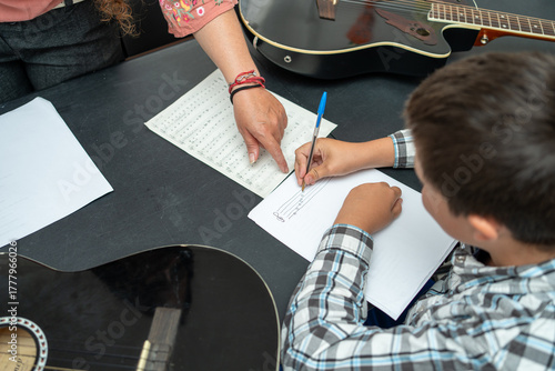 Child writing music notes beside guitar teacher during a private lesson. Music education scene emphasizing composition, theory learning, and how to read and write sheet music effectively together