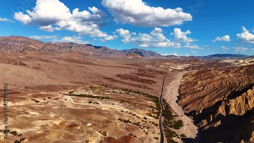 Flight over the highway along the dry desert and bare rocks. White fluffy clouds hang in the blue sky above Death Valley, California, United States.