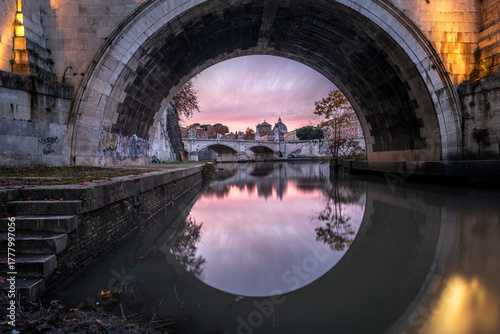 Fototapeta Naklejka Na Ścianę i Meble -  Cityscape in Rome with St Peter's basilica framed by the arch of Sant'Angelo bridge and reflected in the Tiber river at the sunset, Rome, Italy
