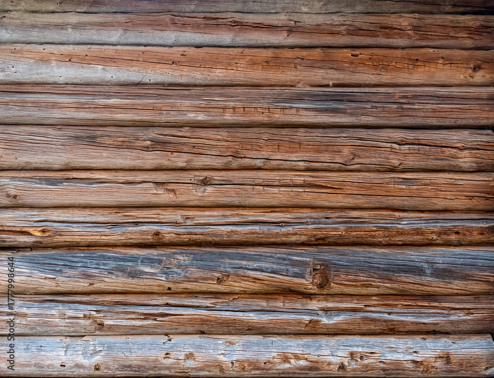 Obraz premium close-up view of a wall constructed from weathered, horizontal wooden logs. variety of textures and natural color variations, with some appearing aged and greyed, while others retain a warm brown hue.