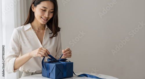 asian woman wrapping elegant blue gift box with ribbon at home during holiday season