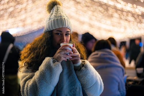 Beautiful woman in a cozy winter outfit enjoys a hot drink at an outdoor festive market with twinkling lights.