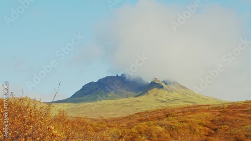 A scenic view of Kristinartindar mountain in Skaftafell National Park, Iceland, with a cloud partially obscuring the peak. The foreground is filled with vibrant orange and yellow autumn foliage under 