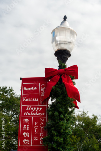 A retro-style tall black metal street lamppost with a bright red cloth bow and a red and white banner hanging from an arm. The words Hanukkah, Kwanzaa, happy holidays, feliz navidad, and Christmas.