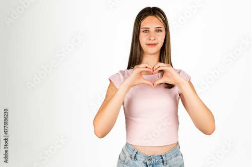 Teenage girl with long hair in a pink shirt and jeans making a heart shape with her hands, isolated on white background. Suitable for concepts of love, kindness, youth, and positive emotions in