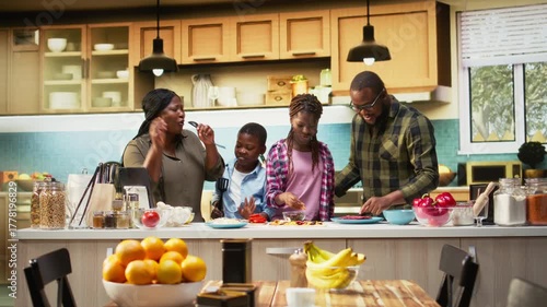 African american young family singing and dancing around in the kitchen, acting silly and laughing together for fun weekend activity. Goofy kids and parents using utensils for karaoke. Camera A.
