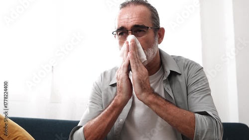 Elderly man sitting on sofa sick using tissue paper sneezing coughing feeling unwell having cold at home. Health concept.

