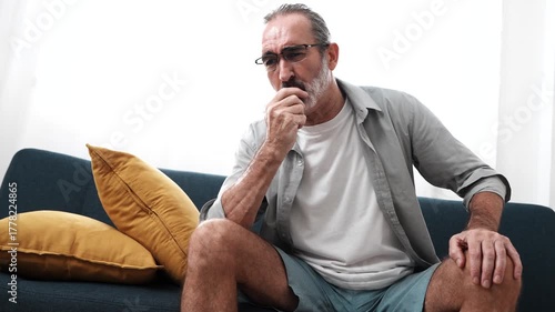 Elderly man sitting on sofa sick using tissue paper sneezing coughing feeling unwell having cold at home. Health concept.

