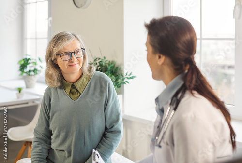 Positive elderly female patient consulting with a doctor or nurse in a hospital office during an appointment. Medical care, healthcare services, and relief provided to patients in need.