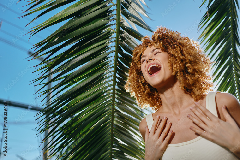 Fototapeta premium Laughing woman with curly hair enjoys sunny day outdoors near lush palm leaves, expressing happiness and natural beauty under clear blue sky.