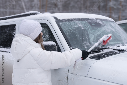 A woman in a white jacket cleans snow and ice with a brush from the windshield of a white car in winter, a close-up view from the back. 