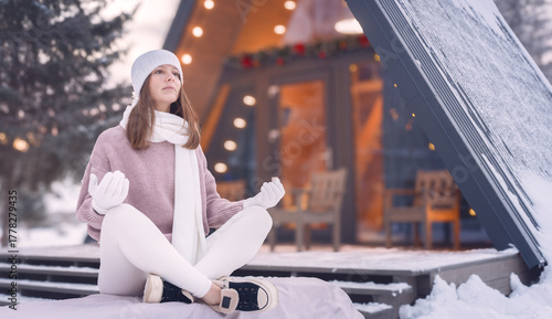 Meditating woman in knitted winter attire by her triangular A-frame wooden house amidst a snowy forest