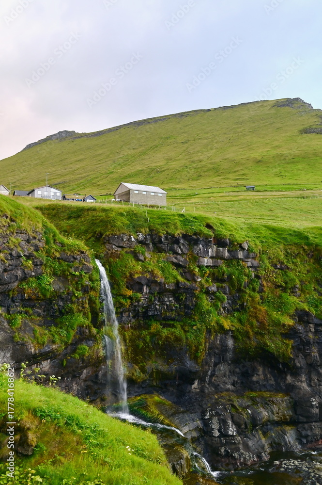 Fototapeta premium Picturesque Village with Waterfall Flowing into the Ocean, Surrounded by Steep Cliffs and Green Hills in the Faroe Islands. Iconic Nordic Landscape