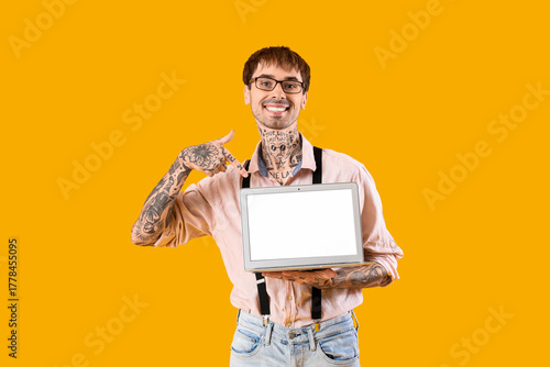 Happy young man pointing at blank screen of modern laptop on yellow background