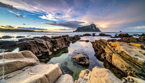 Fototapeta Naklejka Na Ścianę i Meble -  Rocky Shoreline With Footprints Leading To Calm Ocean Water Under A Dramatic Cloudy Sunset Sky With Distant Island Silhouette