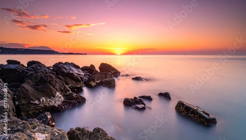 Serene Rocky Shoreline At Sunrise With Soft Orange And Purple Hues Reflecting On Calm Water And Distant Hills Under A Cloudy Sky