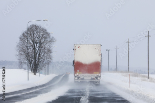 Road landscape of a semi truck driving along highway in snowfall on a day of winter. Difficult road conditions. Rear view, copy space.