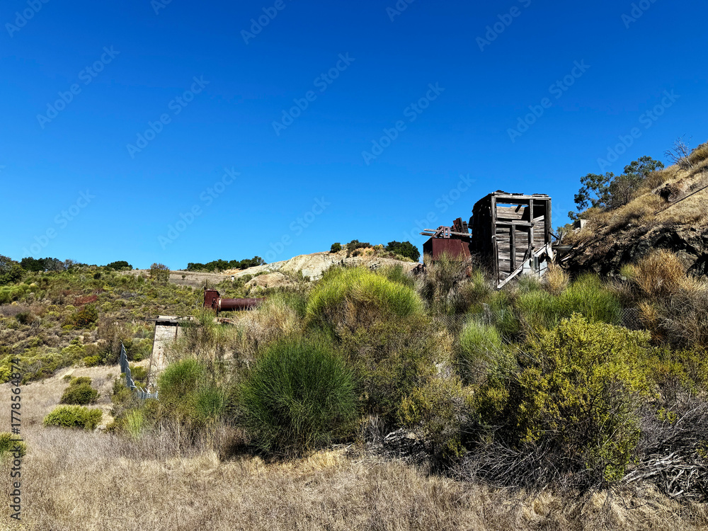 Fototapeta premium Historic rotary furnace ruins in Almaden Quicksilver County Park surrounded by dry grassland, shrubs, and rolling hills under a clear blue sky on a sunny day