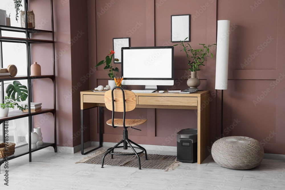 Fototapeta premium Interior of modern office with workplace, shelving unit and floor lamp near brown wall